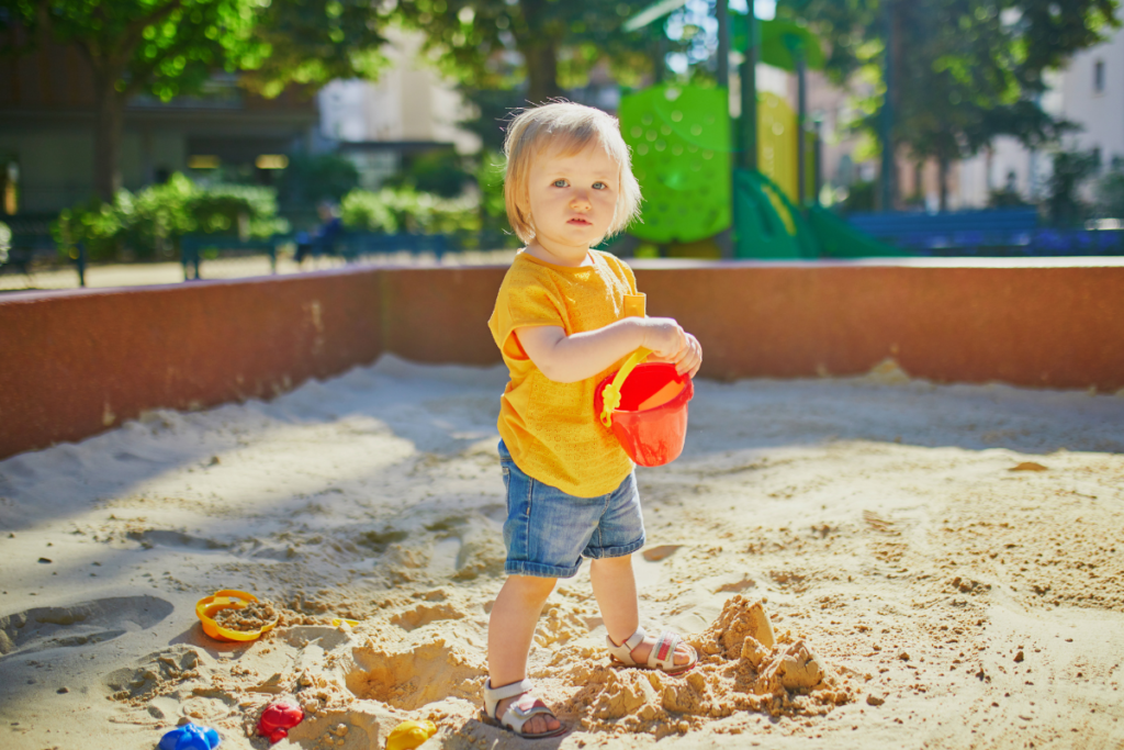 J&oacute;l szeretni.
Egy kisgyermek &aacute;ll a homokoz&oacute;ban, kez&eacute;ben piros v&ouml;d&ouml;rrel