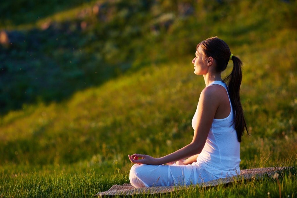 Nő f&eacute;lguggol&oacute; medit&aacute;ci&oacute;s testtart&aacute;sban &uuml;l a fűben, naplement&eacute;ben